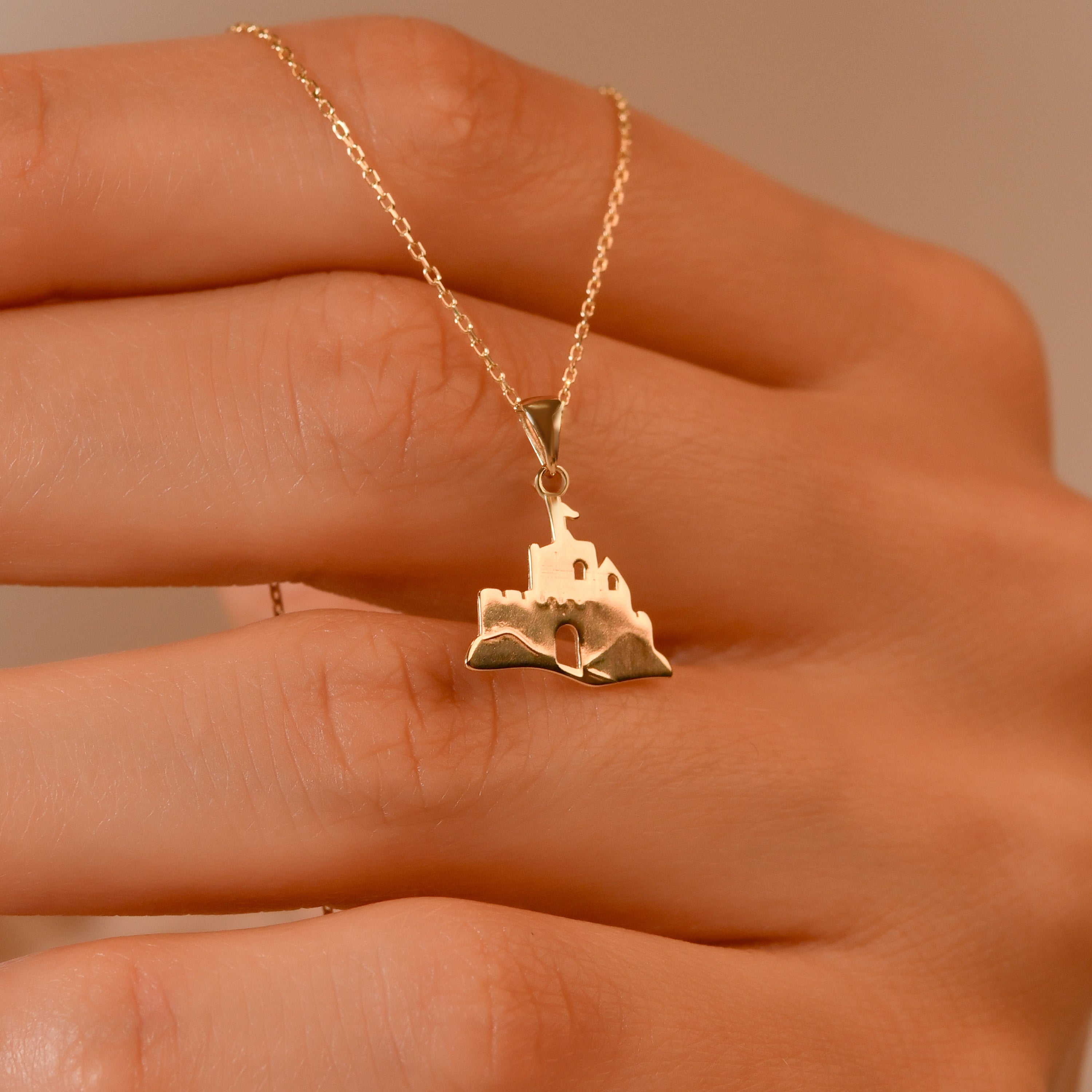 A gold necklace with a small castle-shaped pendant is shown resting on a person's hand.