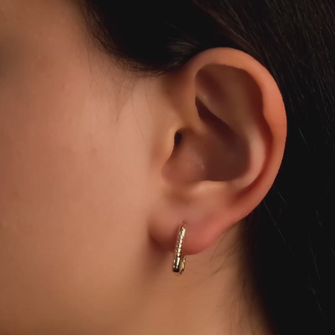 Close-up video of a woman's ear wearing a pair of 14ct solid gold rectangular hoop earrings with ribbed detailing.