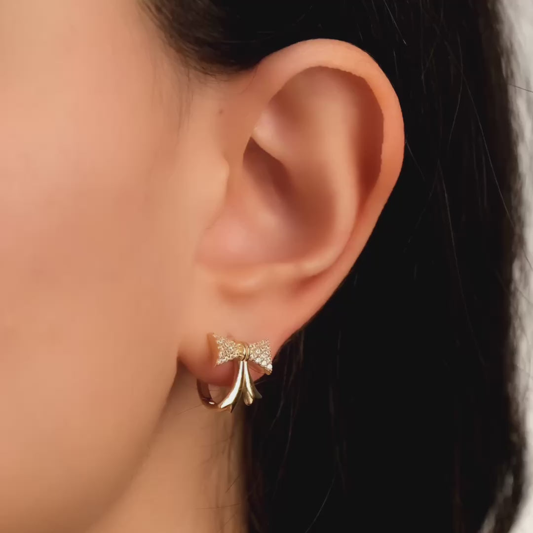 Close-up video of a model wearing solid gold hoop earrings with a charming bow design embellished with cubic zirconia stones, showcasing a fun and stylish accessory.