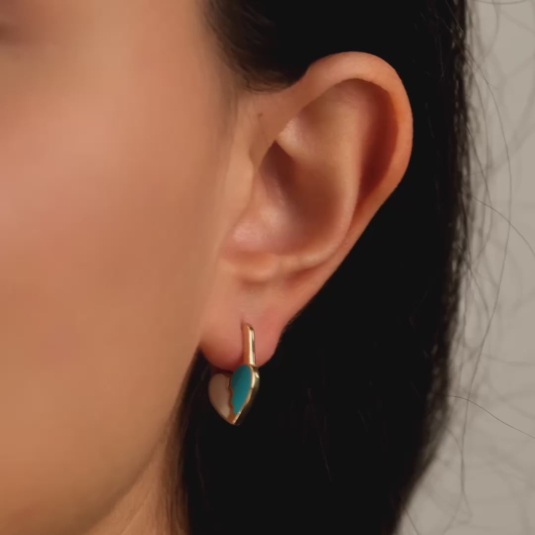 Close-up video of a model wearing solid gold heart-shaped earrings with a broken heart design, showcasing one half in turquoise enamel and the other in a light cream colour.